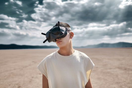 A young person wearing large FPV goggles stands in a vast desert under a dramatic, cloudy sky, blending technology with a remote natural setting.